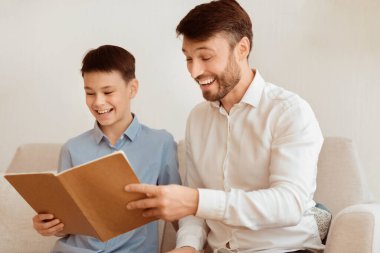 Mutlu baba ve oğul kitap okuyor ve evdeki koltukta birlikte gülüyorlar. Ebeveynlik ve aile ilişkisi. Seçici Odaklanma
