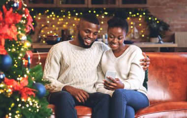 Akıllı telefon kullanarak gülümseyen Afrikalı bir çiftin portresi. Yılbaşı dileklerini okurken evdeki Noel ağacının yanındaki kanepede otururken..
