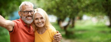 Neşeli yaşlı bir çift birbirine yakın durur, geniş bir gülümsemeyle yeşil ağaçlar ve yumuşak otlarla dolu canlı bir parkta selfie çeker ve kucaklaşırlar. Güneş ışıl ışıl parlıyor, boşluğu kopyala