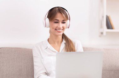 Çevrimiçi Eğitim. Dizüstü bilgisayar kullanan, kulaklıkla podcast dinleyen evde koltukta oturan gülümseyen kız. Seçici Odaklanma