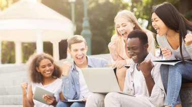 Bir grup çok ırklı öğrenci başarıyı kutluyor, sınav sonuçlarını internetten kontrol ediyor, parkta vakit geçiriyor.