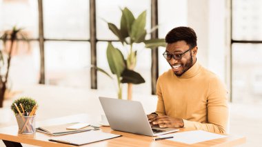 Heyecanlı Adam. Gözlüklü mutlu siyah adam sabah erkenden işlerini halletmek için dizüstü bilgisayarla çalışıyor.