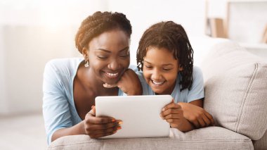 Afrikalı anne ve kızı dijital tablette çizgi film izliyor, kanepeye uzanıyor. Boş alan