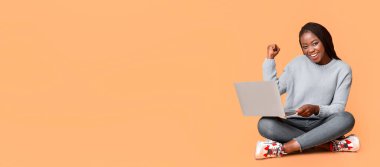 Genç siyah bir kadın dizüstü bilgisayarında başarılı bir görevi tamamladıktan sonra sevinçle kutluyor. Rahat bir kazak giyiyor ve yüzünde parlak bir gülümseme var.