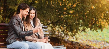 Neşeli adam kız arkadaşına yeni cep telefonu uygulamasını gösteriyor, halka açık bir parkta randevusu varken, bankta oturuyor, kahve içiyor ve akıllı telefon ekranına bakıyor, boş alan arıyor.