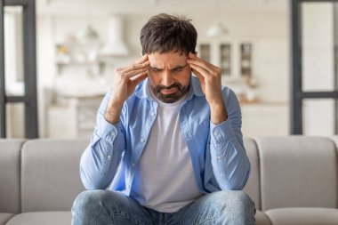 Oturma odasındaki bir kanepede oturan bir adam başını tutuyor ve baş ağrısı çekiyor gibi görünüyor. Mavi bir gömlek ve kot pantolon giyiyor..
