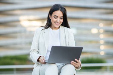 Çağdaş mimariyle çevrili bir ortamda, takım elbiseli genç bir kadın bilgisayarını kullanırken gülümsüyor..