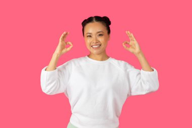 Neşeli genç bir Asyalı kadın parlak pembe bir arka planda el işareti yapıyor..