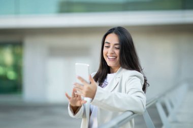 Bir kadın, modern bir mimari ortamda ekranda gülümseyen akıllı telefonunu açık havada kullanmaktan zevk alır..