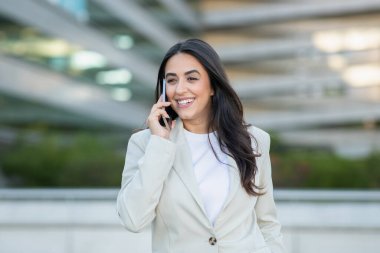 Bir kadın akıllı telefonuyla konuşurken gülümsüyor, açık renk ceket giymiş, çağdaş bir şehir zeminine karşı..