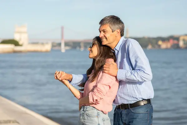 stock image A couple stands together by the water in Lisbon, sharing a warm embrace and laughter. The vibrant city backdrop and gentle waves create a picturesque setting on a sunny day.