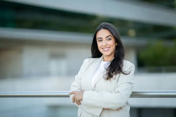 stock image A young woman stands confidently with her arms crossed, smiling warmly, in an urban area with modern architecture.