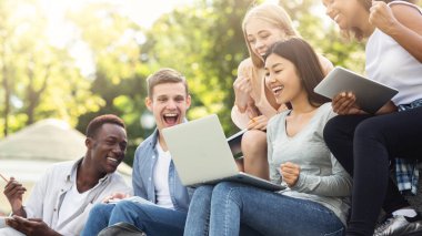 Group of emotional multiethnic students celebrating success, checking exam results online in park, copy space