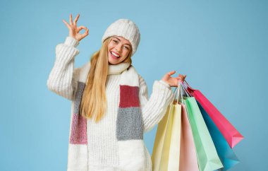 Sıcak kış kıyafetleri içinde gülümseyen güzel bir kız tamam işareti, alışveriş torbaları, sarı arka plan.