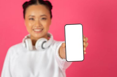 Genç bir Asyalı kadın elinde boş bir akıllı telefonla, kulaklık takarak ve parlak pembe bir arka planın önünde durarak gülümsüyor..