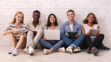 Diz üstü bilgisayar, kitap, dijital tablet ve panorama kullanarak beyaz tuğla duvarın üzerinde okuyan neşeli uluslararası öğrencilerden oluşan bir grup.