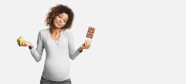 Kafası karışmış Afro-Amerikalı hamile kız, elinde elma ve çikolata tutuyor, ne seçeceğini bilmiyor, beyaz stüdyo geçmişi var.