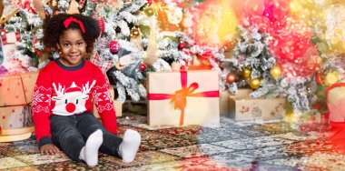 Güzel Afrikalı Amerikalı kız Noel ağacının yanında poz veriyor, dekore edilmiş oturma odasında oturuyor ve kameraya gülümsüyor.