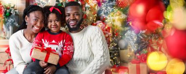 Üç kişilik neşeli afro aile Noel ağacı arka planında hediyeyle poz veriyor, gülümsüyor ve kameraya bakıyor, yakın plan