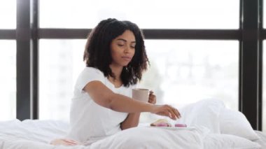 Genç Afro-Amerikan bayan yatakta kahvaltı yapıyor, sabahları kahveyle kurabiye yiyor, yavaş çekimde.