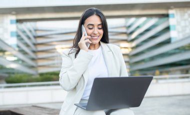 Bilgisayarını kullanarak bir telefon görüşmesi yapan bir kadın, modern mimariyle çevrili bir çalışma alanında gülümsüyor..