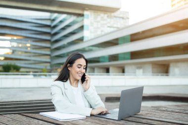 Bir kadın, şehirdeki modern bir çalışma alanında dizüstü bilgisayarıyla yazı yazarken bir telefon görüşmesi yapıyor..