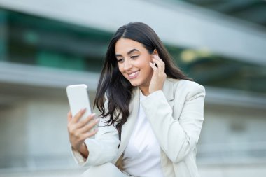 Hafif takım elbiseli genç bir kadın modern bir binanın dışında telefonuyla mutlu bir şekilde meşgul oluyor.