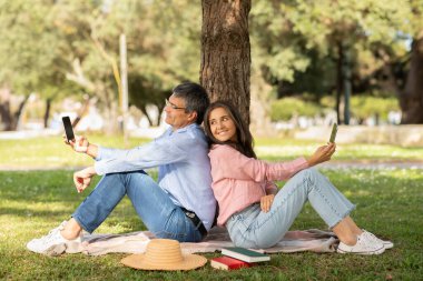 Bir çift parkta bir battaniyenin üzerinde sırt sırta oturur. Güneşli bir günde yeşilliklerle çevriliyken akıllı telefonlarının keyfini çıkarıyorlar, rahat ve rahat bir atmosfer yaratıyorlar..