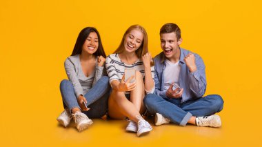 Gambling online. Happy international friends celebrating success, looking at mobile phone screen, yellow background, panorama