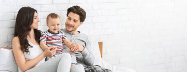 Neşeli genç bir çift bir yatakta oturur, neşeli bebekleriyle meşgul olurlar. Ebeveynler birbirlerine gülümserler ve çocuk sıcak bir atmosfer yaratır. Yumuşak ışık odayı dolduruyor, boşluğu kopyalıyor