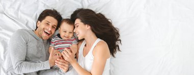 Mutlu bir aile, yatağında hassas bir anın tadını çıkarıyor. Ebeveynler gülümsüyor ve küçük çocuklarıyla etkileşime geçiyorlar.