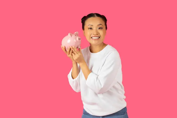 stock image A happy Asian woman smiles while holding a pink piggy bank in front of a vibrant pink backdrop.