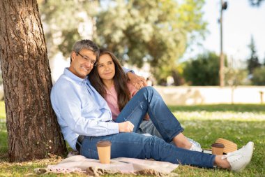 Bir çift, yemyeşil bir parkta, bir ağaca yaslanmış, battaniyenin üzerinde birlikte oturuyorlar. Sıcak bir anı paylaşıyorlar, kahveyi yudumlarken gülümsüyorlar, güneşli günün tadını çıkarıyorlar..