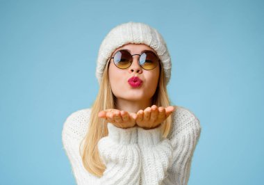 Sarı arka planda güneş gözlüğü ve kış şapkası takan genç, güzel sarışın kadın, havada el sallayan öpücük, aşk ifadesi.