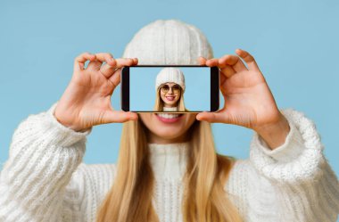 Güzel bir kış kadını yüzünü akıllı telefonla kapatıyor. Selfie 'sini ekranda gösteriyor. Stüdyonun sarı arka planı.