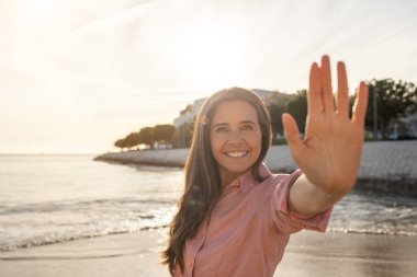Bir kadın kıyıda duruyor, gülümsüyor ve elini sallıyor güneş suyun üzerinde yükselirken, sakin bir atmosfer yaratıyor. Sahil sakin, yumuşak dalgalar ve hafif ışıklarla dolu..