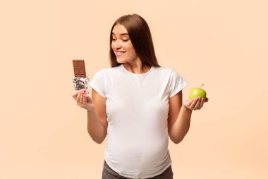 Gebelik Diyet ve Beslenme. Hamile Kadın Beyaz Stüdyo Arka Plan Üzerinde Duran Elma ve Çikolata Arasında Seçim.
