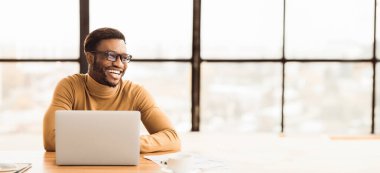 Başarılı bir Freelancer. Mutlu siyahi adam kopyalama alanına bakıyor, modern dizüstü bilgisayar kullanarak ev ofisinde çalışıyor.