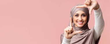 Güzelliğini çerçevele. tesettürlü gülümseyen müslüman kız parmaklarıyla çerçeve yapıyor, anı yakalıyor, pembe arkaplanda boş alan ile poz veriyor