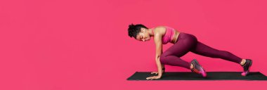 Black fitness girl working out over red studio background, exercising on fitness mat, listening to music with wireless earphones, panorama with free space