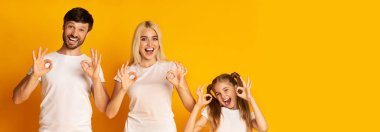 Bir baba, bir anne ve bir kızdan oluşan neşeli bir aile bir arada duruyor, herkes gülümsüyor ve iyi bir el hareketi yapıyor. Şakacı ifadeleri ve uyumlu kıyafetleri.