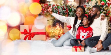 Şenlikli selfie. Mutlu afro kadın kocası ve kızıyla Noel ağacı ve şöminenin yanındaki dekore edilmiş oturma odasında fotoğraf çekiyor.