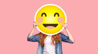 Kızıl saçlı kız, gülümseyen emoji gülüşünün arkasına saklanıyor, pembe stüdyo arka planında poz veriyor, taklit alanı olan bir panorama.