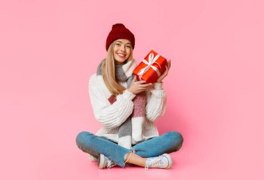 Kışlık giysiler içinde gülümseyen kadın yerde oturuyor, elinde Noel hediyesi, sarı stüdyo geçmişi var.