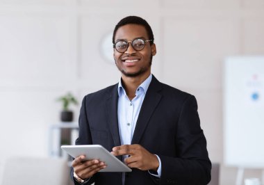 Elinde dijital tablet olan neşeli, genç siyahi bir menajer. Kameraya gülümsüyor. Ofis içi, fotokopi odası. Afrikalı Amerikalı işadamı iş başvurusunu ped üzerinde kullanıyor