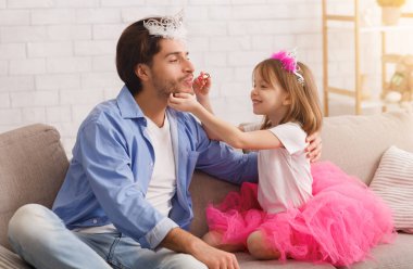 Küçük tatlı bir kız ruj sürüyor baba dudaklarına, baba ve çocuk taçlı, ev içi.