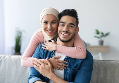 Evlerinde kucaklaşan neşeli Müslüman bir çiftin portresi, fotokopi alanı, yakın çekim. Gülümseyen genç bir kadın, eşarplı sevgili kocasına arkadan sarılıyor, Orta Doğulu bir aile birlikte vakit geçiriyor.