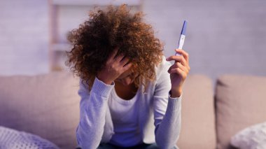 Depresyondaki kız hamilelik testi tutuyor. Yüzü elle kaplanmış, koltukta oturuyor, boş yer var.