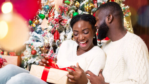 Радостная африканская американка получает рождественский подарок от своего заботливого мужа, сидя дома возле елки
