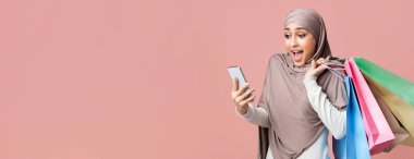 Mevsimlik satışlar. Akıllı telefon kullanan ve pembe arka planda açık alan olan parlak alışveriş torbalarını tutan tesettürlü Arap kız şok oldu.